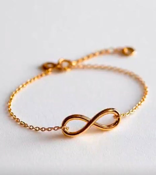 A delicate gold chain bracelet featuring a polished infinity symbol centerpiece, displayed on a clean, light off-white background.